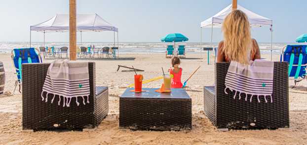 Two chairs on the beach are seen from the back. A blonde women sits in one chair and a table with tropical drinks sits between the chairs. A child plays in the sand in front of the setup.