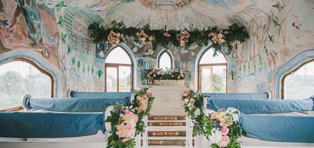 The interior of a chapel painted with murals is decorated with pink, white, and green florals, and white pews are draped in light blue fabric
