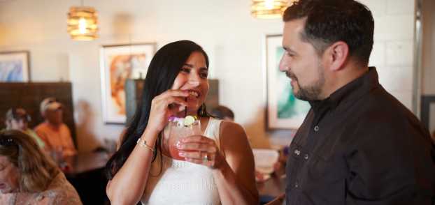 A woman with long hair sips on a cocktail and smiles at the man next to her