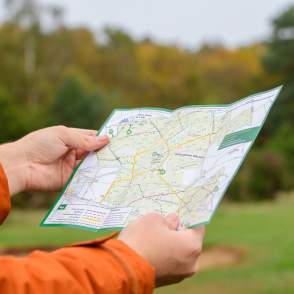 a close up of a man holding a walking guide map with a bright orange coat on and autumnal trees in background