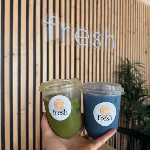 Hand holding up two Fresh juice drinks in front of a logoed wall.