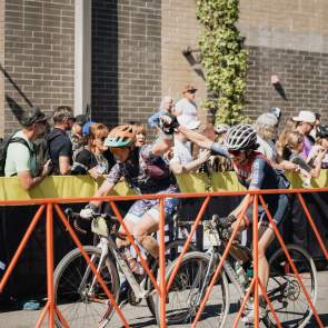 Two cyclists with raised arms hold hands in celebration as they approach the finish line in Bentonville, Arkansas as spectators cheer.