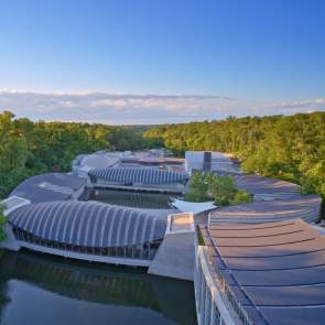 Aerial view of modern architectural structures nestled among lush greenery, reflecting on a serene waterway under a clear blue sky.
