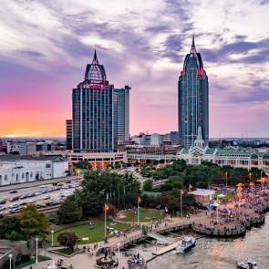A vibrant sunset over Downtown Mobile skyline, featuring tall buildings, a bustling waterfront, and a lively crowd below.