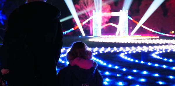 A mother and small child stand with their backs to the camera looking at a small, lit up London Bridge. There are roaming spotlights & glowing fairylights on the floor in all different colours. The trees off in the distance are also lit up in blue and pink.