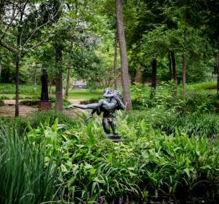 Bronze sculpture of one person holding another amidst lush greenery in an outdoor sculpture garden.