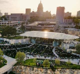 Aerial view of Moody Amphitheater in evening sun. There is a crowd on the open lawn and in seats near the pavilion stage. The State Capitol building is visible behind the amphitheater