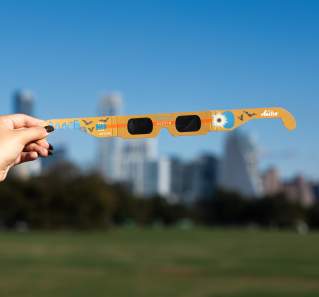 Woman's hand holding a pair of Austin eclipse glasses in front of the downtown skyline.