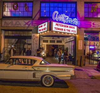 People buzzing in and out of Antone's at night. The iconic neon sign is lit and a vintage car is parked out front of the venue.