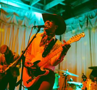 Musician in a cowboy hat singing into a mic while playing guitar with colorful lights surrounding him.