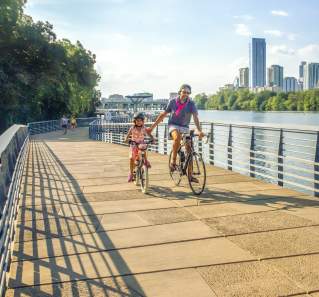 Image of a man and daughter riding bikes on the Butler Hike and Bike trail boardwalk with Austin Downtown in the background.