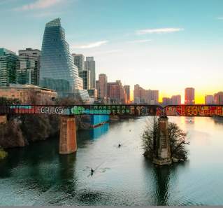 Kayakers paddling under a bridge with grafitti on Lady Bird Lake while the sun sets against Downtown Austin buildings.