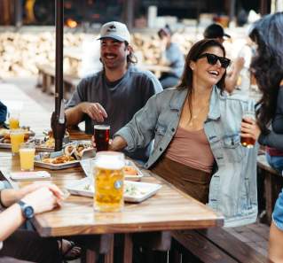 Group of friends sitting, laughing together at a picnic table with steins of beer on a sunny day.