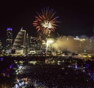 Red fireworks explodes over downtown Austin skyline and Lady Bird Lake at night. Aerial view shows a music stage and crowd gathered below at Auditorium shores.