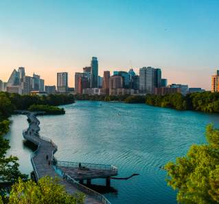 People walking along suspended board walk that follows the shape of the Colorado River, leading to Downtown Austin skyline.