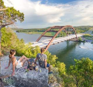 Hikers sitting on a hilltop rock overlooking the Pennybacker Bridge at Lake Austin.