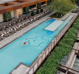 Aerial photo of the JW Marriott rooftop pool with a woman in a red bathing suit swimming on her back in the center of the pool. The floor of the pool includes an outline of the state of Texas with a star on the location of Austin.