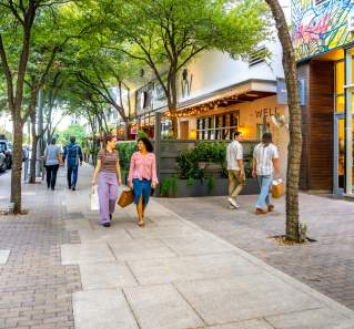 Two women walking down the sidewalk under trees on 2ND Street District with shopping bags