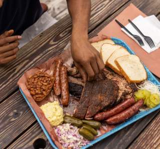 Plastic tray covered in butcher paper topped with slices of different barbecue meats and traditional sides.