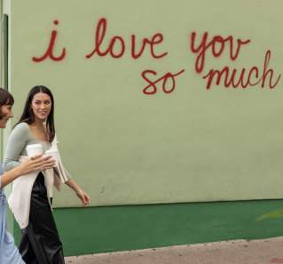 Two women walking in front of the I Love You So Much mural with coffee in hand.