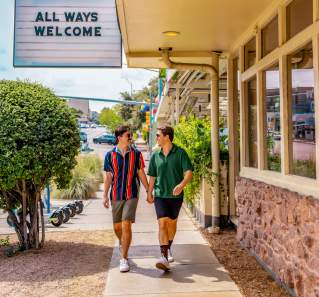 Two men walk down the sidewalk in front of Joann's Fine Foods, holding hands under a marquee sign reading "All Ways Welcome" with a neon pair of lips at the top.