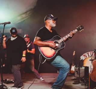 Musician Ray Prim plays an acoustic sunburst guitar on stage at Stubb's in Austin Texas. Two other male musicians stand behind him