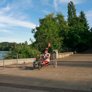 A person on a three-wheeled recumbent bike rolls down the wide smooth paved path alongside the Willamette River. The cyclist is wearing warm weather clothing. The sky is blue and the trees are bright green.