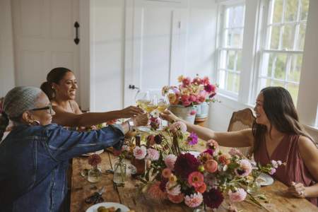 Three women raise glasses of white wine in a toast across a flower-covered farm table inside a bright Loudoun County venue.