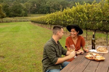 A couple enjoying a picnic and a bottle of wine on a picnic table next to grape vines