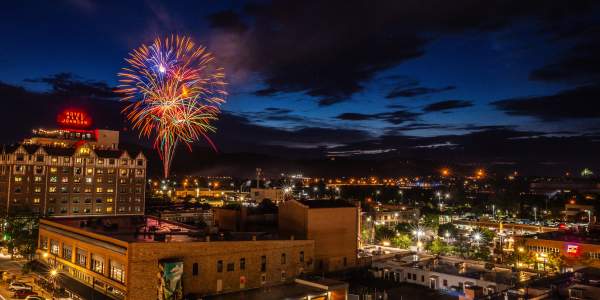 Fireworks burst in the night sky above downtown Rapid City, with the illuminated Hotel Alex Johnson and city lights glowing beneath dark clouds at dusk.