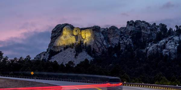 Mount Rushmore, lit up by yellow floodlights at dusk, sits in the background as a vehicle traveling down the highway is trailed by a long line of red taillights, created by a long-exposure photo.