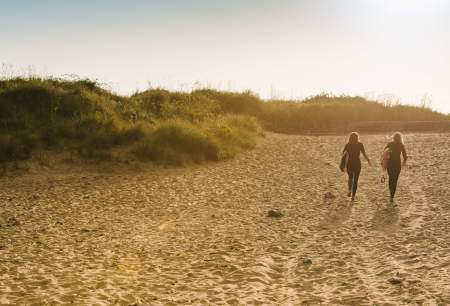 Two ladies walking across the beach with surfboards