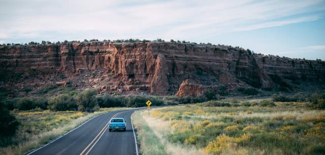 A car drives on a winding road past a large red rock formation under a clear blue sky.