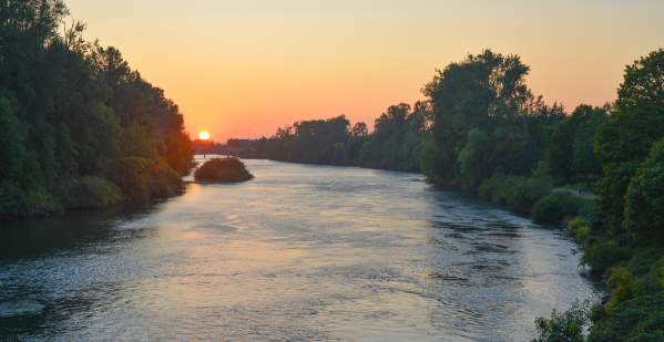 The sun sets on the Willamette River. A wide river bordered by green trees and the sky is orange and red.