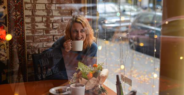 looking through a window at a woman smiling with a mug of coffee. there are flowers on her table and fairy lights in the background.