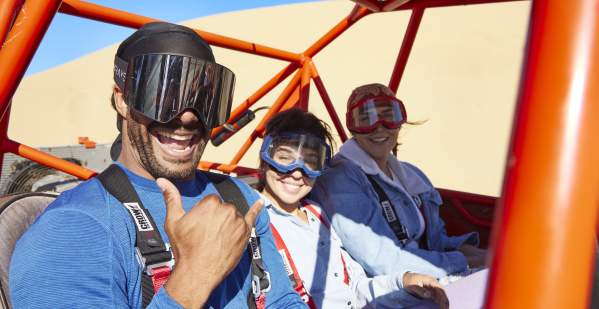 Dune buggy riders in goggles, smiling and throwing a Shaka.