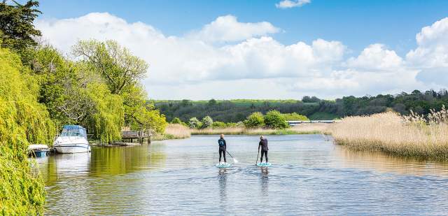 Two paddle boarders on the River Arun, with a boat moored to the left and grassy bank to the right, in Sussex