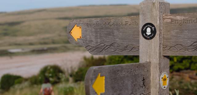 A wooden sign post with yellow arrows pointing to England coast path, with a walker in the distance