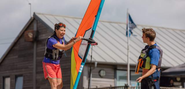 Windsurf instructor looks at pupil who is standing on a windsurf with orange sail, on Eastbourne Beach