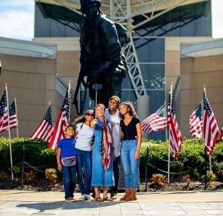 Family posing together in front of a large military statue with American flags displayed outside a museum building