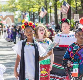 Group of children and teens in vibrant cultural dress participating in the International Folk Festival parade on Hay Street in Fayetteville, NC, with flags waving and crowds watching.