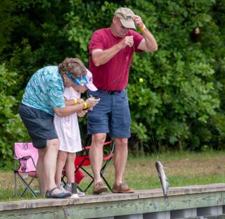 Family participating in a fishing workshop, helping a child reel in a catch from a dock beside the water