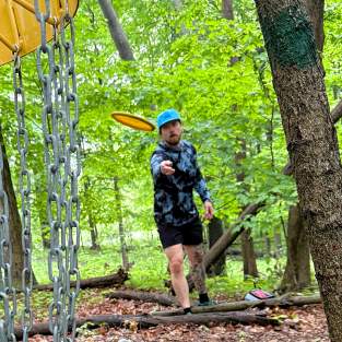 Man throws disc golf frisbee towards camea, which sits behind the hole in heavily wooded and shaded area