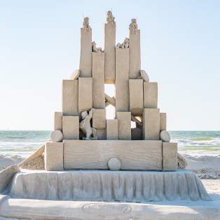 Sand sculpture depicting small men playing instruments and building a structure out of blocks.