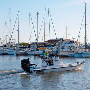 A fishing boat with two people in the driver's seat putters through a marina with rows of sailboats behind