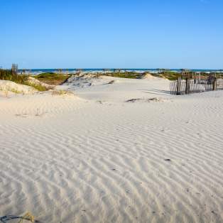 Groomed sand with grassy dunes in the background. Water is visible in the background.
