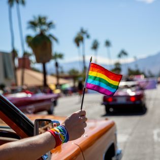 Pride flag waving outside of car in downtown Palm Springs with mountains and palm tree