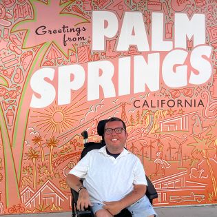 Man in wheelchair smiling for a photo in from of a Palm Springs mural