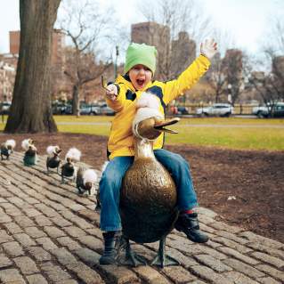 Child sitting on duck statue with line of duckling statues lined up behind it