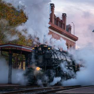 A large black steam train engine pulls into the Cumberland Station as it releases a billowy cloud of steam.